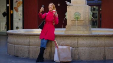 Young attractive woman with shopping bags in an old city taking a selfie. Long hair female in red coat is holding a takeaway coffee cup near an antique fountain. Slow motion 4k video. 