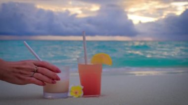 Close up of cocktails at sunset. Hand of a woman taking away a drink. Two drinks stand. Blue turquoise ocean on the background. Summer travel vacation concept. 4k slow motion video. 