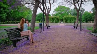 Kadın telefonuyla selfie çekiyor. Mor jakarandası olan güzel bir park. Romantik büyülü atmosfer. Yeşil yemyeşil yapraklar. Yaz doğası.