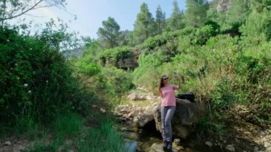 Kadın bir taşın üzerinde oturuyor ve selfie çekiyor. Küçük gölü olan güzel bir manzara. Yeşil gölet ve taşlar. Güneşli güzel hava. Mavi gökyüzü. Yatay video. 