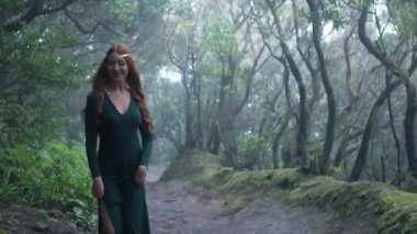 Beautiful woman in an elf costume is walking in a magic green enchanted forest. Redhead girl with a long hair. Trees covered with most on the background. 