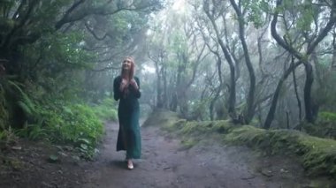 Beautiful woman in an elf costume is walking in a magic green enchanted forest. Redhead girl with a long hair. Trees covered with most on the background. 