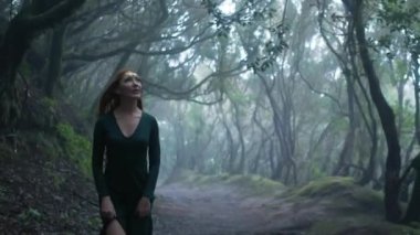 Beautiful woman in an elf costume is walking in a magic green enchanted forest. Redhead girl with a long hair. Trees covered with most on the background. 