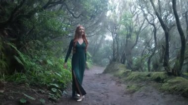 Beautiful woman in an elf costume is walking in a magic green enchanted forest. Redhead girl with a long hair. Trees covered with most on the background. 