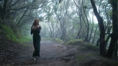 Beautiful woman in an elf costume is walking in a magic green enchanted forest. Redhead girl with a long hair. Trees covered with most on the background. 