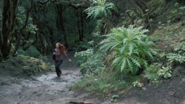 Woman is running in an enchanted forest. Redhead girl with a long hair holding a backpack. Trees covered with most on the background. 