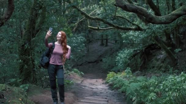 女は魅惑的な森の中で自撮りをしている 携帯電話を持っている長い髪を持つ赤毛の女の子 背景に最も覆われた木 — ストック動画