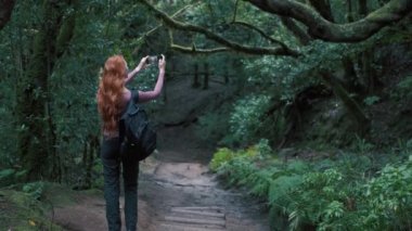 Woman is in an enchanted forest taking a selfie. Redhead girl with a long hair holding a mobile phone. Trees covered with most on the background. 