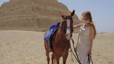Beyaz elbiseli kadın ayakta ve bir atla gülümsüyor. Arka planda Djoser Step var. Arkeolojik olarak Saqqara mezarlığı, Mısır, Afrika. Seyahat konsepti.