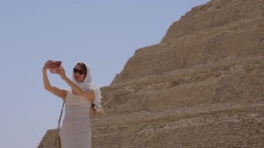 Beyaz elbiseli kadın selfie çekiyor. Arka planda Djoser Step var. Arkeolojik olarak Saqqara mezarlığı, Mısır, Afrika. Seyahat konsepti.