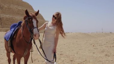 Beyaz elbiseli kadın ayakta ve bir atla gülümsüyor. Arka planda Djoser Step var. Arkeolojik olarak Saqqara mezarlığı, Mısır, Afrika. Seyahat konsepti.
