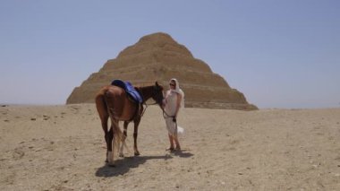 Beyaz elbiseli kadın ayakta ve bir atla gülümsüyor. Arka planda Djoser Step var. Arkeolojik olarak Saqqara mezarlığı, Mısır, Afrika. Seyahat konsepti.