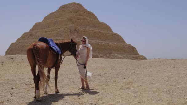 穿着白衣的女人站在那里 和马一起笑着 背景是Djoser Step 考古遗存于埃及 非洲的萨卡拉墓地 旅行旅游概念 — 图库视频影像