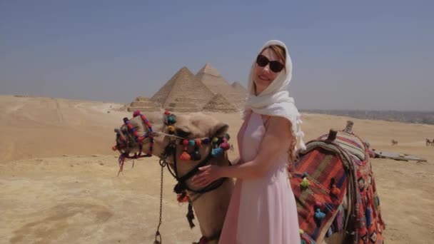 若い女性はエジプトのラクダの近くに立っている ギザの大ピラミッドが背景にあります 女の子は幸せから彼女の腕を上げている — ストック動画