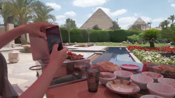 埃及菜在桌子上 阿拉伯菜 吉萨金字塔在背景上 富丽堂皇的酒店露台 有棕榈树 还有一座宏伟的金字塔 拍照的女人 — 图库视频影像