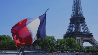 Mavi arka planda Fransız bayrağı olan Eyfel Kulesi. Arka planda yeşil ağaçlar ve Seine nehri var. Yavaş çekim videosu.
