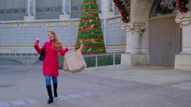 Alışveriş torbaları ve bir fincan kahveyle kırmızı ceketli mutlu bir kadın. Arka planda parlak ışıkları olan Noel ağacı var. Tatil konsepti. Yavaş çekim videosu. 