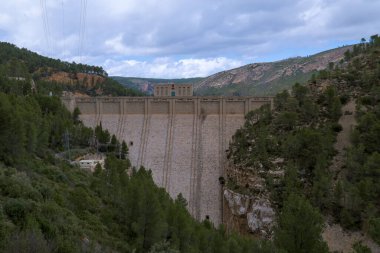 Görüntü, iki orman tepesi arasında yer alan büyük bir baraj yapısını gösteriyor. Baraj betondan yapılmıştır ve tepesinde bir kontrol binası vardır. Çevresindeki manzara yeşilliklerle doludur..