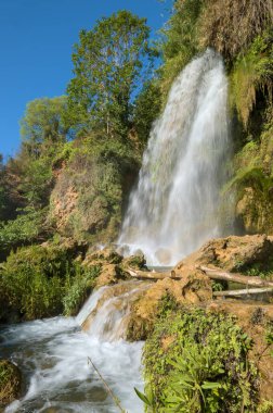 Resim, yemyeşil bitki örtüsüyle çevrili kayalık bir uçurumdan çağlayan bir şelaleyi gösteriyor. Temiz su, temeli parlak mavi gökyüzü olan bir dereye akar..