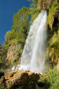 Görüntü kayalık bir uçurumdan aşağı çağlayan güçlü bir şelaleyi ve engebeli arazide hızla akan suyu gösteriyor. Gökyüzü parçalı bulutlu, dramatik doğal manzaraya ekleniyor.