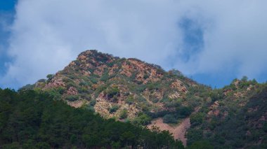 Kayalık çıkıntılı yemyeşil bir dağ sırası parçalı bulutlu bir gökyüzünün altında tasvir edilir. Yoğun bitki örtüsü ve engebeli arazinin birleşimi doğal ve manzaralı bir manzara yaratır..