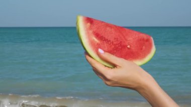 Sahilde bir karpuz dilimini tutan bir el. Arka planda okyanus var. Uzaklaşıyorum. Yaz tatili. Yavaş çekim 4K video.