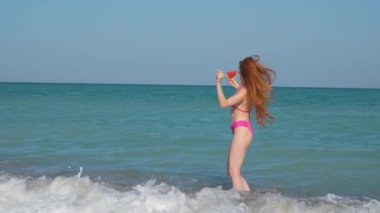 Bir kadın elinde karpuz tutarak mutlu bir şekilde döner ve gülümser. Bu bir tatil konsepti, elleri havada, uzun saçlar uçuşuyor, ve doğal güzellik yavaş çekimde 4k video ile çekiliyor..