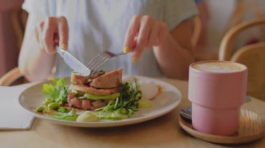 Bu 4K yavaş çekim videosu bir kahvaltı sahnesinin yakın çekimini bir tabakta bir ton balığının elini keserek çeker. Tabağın yanında, latte sanatıyla birlikte pembe bir fincan kahve var..