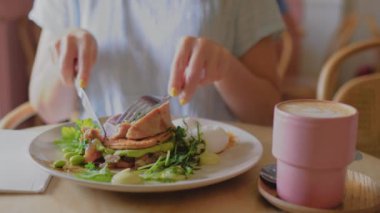 Bu 4K yavaş çekim videosu bir kahvaltı sahnesinin yakın çekimini bir tabakta bir ton balığının elini keserek çeker. Tabağın yanında, latte sanatıyla birlikte pembe bir fincan kahve var..