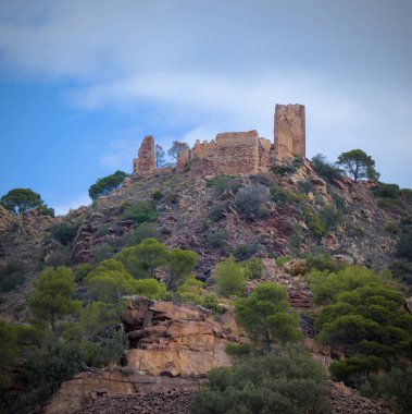 Kayalık bir tepenin üstündeki eski bir kale ya da kalenin kalıntıları. Tepe yeşil çalılar ve ağaçlarla kaplı. Yukarıda, gökyüzü parçalı bulutlu ve mavi lekeler görünüyor..