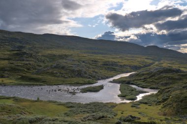 A serene landscape of Norwegian nature featuring rolling hills, a winding river, and lush green vegetation under a dramatic cloudy sky with sunlight breaking through. High quality photo. clipart