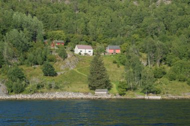 Bu görüntü, sakin mavi bir su kütlesine bakan yoğun yeşil ağaçlarla çevrili, bir kaç renkli evi olan manzaralı bir kır manzarası gösteriyor..