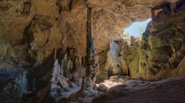 Bu resim doğal bir mağaranın içini gösteriyor, yüksek dikitler, engebeli kaya oluşumları ve büyük bir açıklıktan akan güneş ışığı, toprak dokuları vurguluyor..