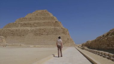 Mavi gökyüzünün altında Djoser Piramidi olarak da bilinen ikonik Djoser Piramidine doğru yürüyen bir turistin yavaş çekim videosu eski Mısır 'ın ebedi cazibesini yakalıyor..