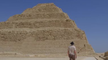 Mavi gökyüzünün altında Djoser Piramidi olarak da bilinen ikonik Djoser Piramidine doğru yürüyen bir turistin yavaş çekim videosu eski Mısır 'ın ebedi cazibesini yakalıyor..