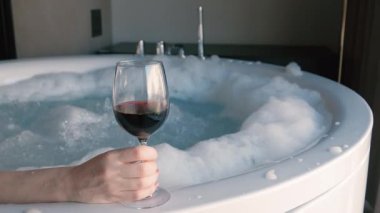 Spa anı, elinde bir bardak kırmızı şarap tutarken köpüren bir jakuzide dinlenen bir insan. Sakinleştirici su, köpük ve zarif ortam rahatlama ve kendine bakmanın özünü vurgular..