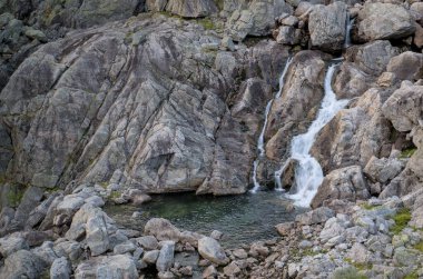 Small cascading waterfall flowing through rugged rock formations into a clear pool, surrounded by a rocky mountain landscape in Norway's pristine natural environment. High quality photo