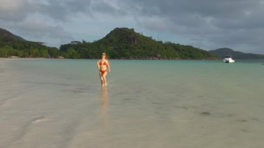 Canlı bir ağır çekim 4K videosu kırmızı bikinili bir kadını, yemyeşil tepeler ve arka planda tropikal tatilinin keyfini çıkararak, çarpıcı bir Seyşeller plajında sığ dalgalarda koşarken yakalıyor..