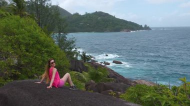 Seyşeller 'de, manzaralı okyanus manzarası ve tropikal manzarası olan bir kayanın üzerinde oturan bir kadının yavaş çekim 4K videosu.. 