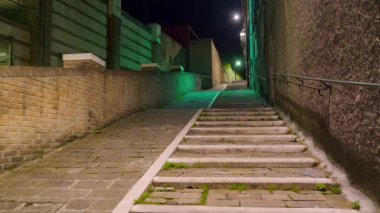 Dik, kaldırımlı bir sokak geceleri yukarıya doğru esiyor yumuşak yeşil ve beyaz ışıklarla aydınlatılan şehir manzarası yolu saran taş duvarlarla dolu. 