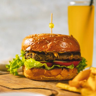 Sulu burger ahşap bir tahtanın üzerinde patates kızartması ve arka planda bir bardak portakal suyu ile duruyor.