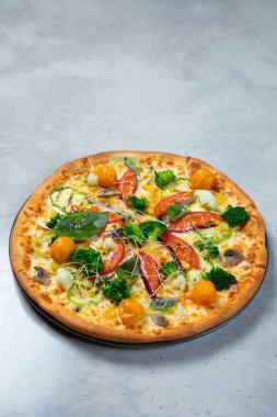 Gri masa arkasında çeşitli sebzeler ve mozzarella peynirli taze pişmiş vejetaryen pizza.