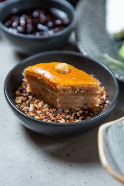 Fındık ve balla kaplı tatlı baklava parçası, siyah kasede ezilmiş fındıkların üzerinde yatıyor.