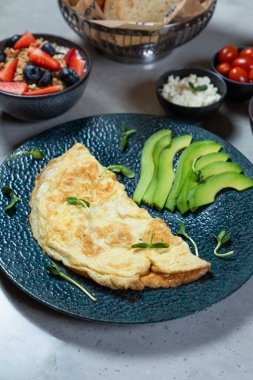 Sağlıklı kahvaltı için mavi tabakta avokado dilimleri ve mikroyeşilliklerle servis edilen omlet.