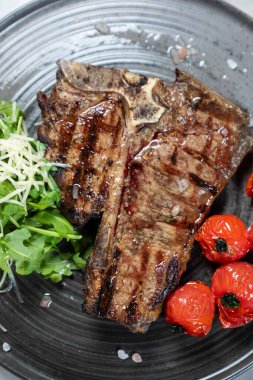 Izgara biftek, ızgara vişneli domates, taze roka ve parmesan peyniri ile servis edilir.