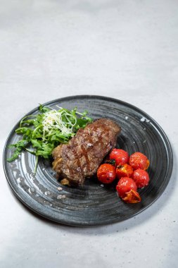 Kızarmış domates ve roka salatasıyla birlikte siyah tabakta servis edilen ızgara biftek görsel olarak çekici ve lezzetli bir mutfak deneyimi yaratıyor.