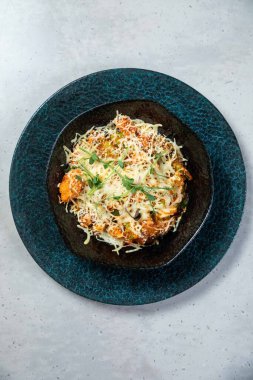 Mavi bir tabakta peynirli ve otlu fırında yemek, yukarıdan hafif bir arka planda çekiliyor.