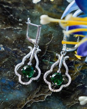 Bir çift zümrüt ve elmas küpe yanında mavi bir çiçekle taş bir yüzeyde duruyor.