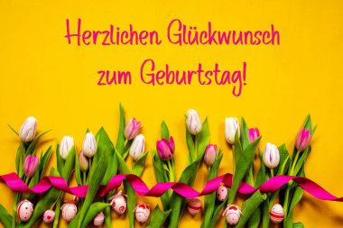 German Text Herzlichen Glueckwunsch Zum Geburtstag Means Happy Birthday. White And Pink Tulip Spring Flowers With Ribbon And Easter Egg Decoration. Yellow Wooden Background