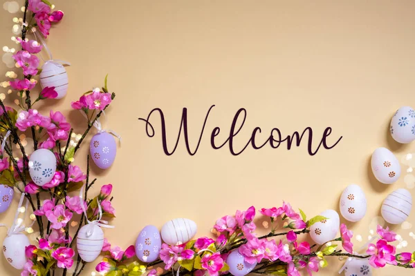 English Text Welcome On Beige Background. Pink And Purple, Shiny And Bright Spring Flower Arrangement With Easter Egg Decoration.